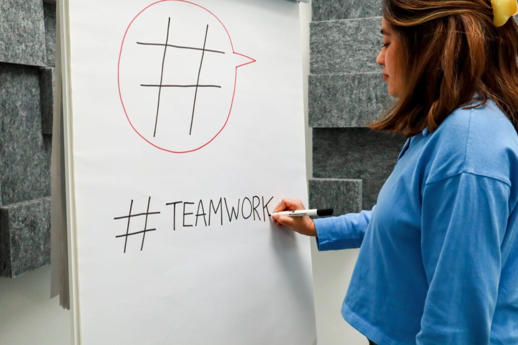 A woman in a jean jacket writes "#TEAMWORK" on a flipchart during a business meeting.
